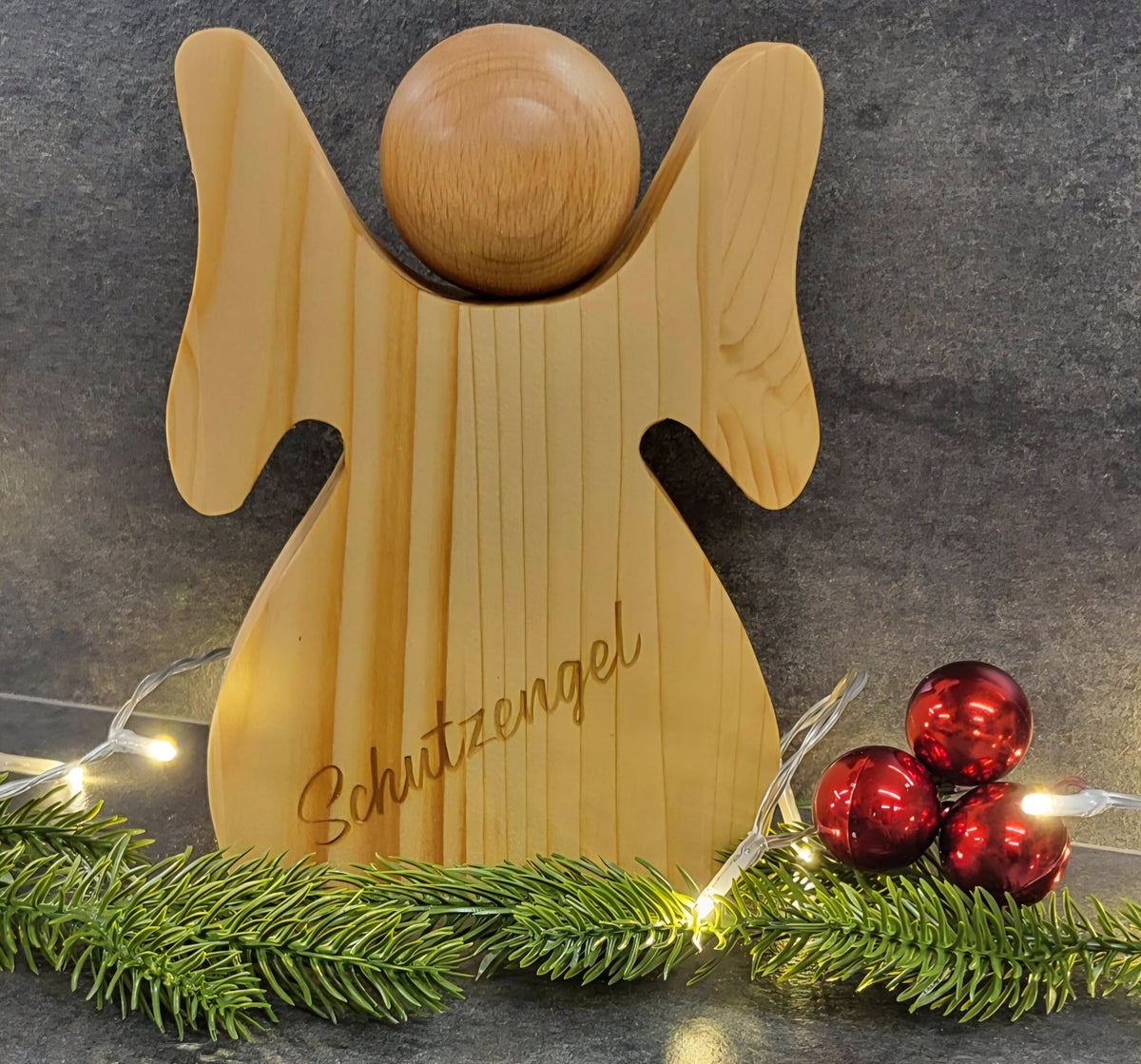 Handgefertigter Schutzengel aus Holz mit individueller Gravur als persönliches Geschenk.
Engel mit Gravur: Schutzengel 
Auf dem Bild sind noch Tannenzweige, Lichterkette und rote Kugeln. Der Engel ist auch ein tolles Weihnachtsgeschenk 
